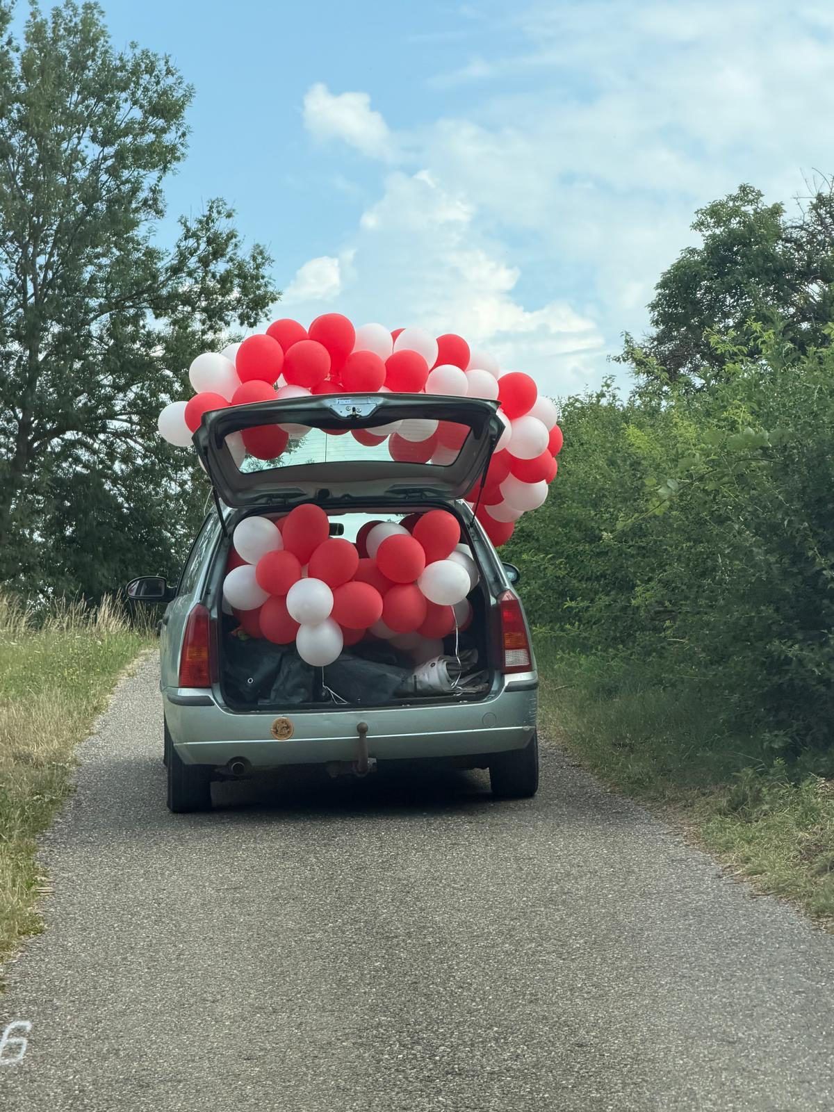 Kombi mit rot-weißer Ballongirlande und offener Heckklappe fährt über einen Feldweg