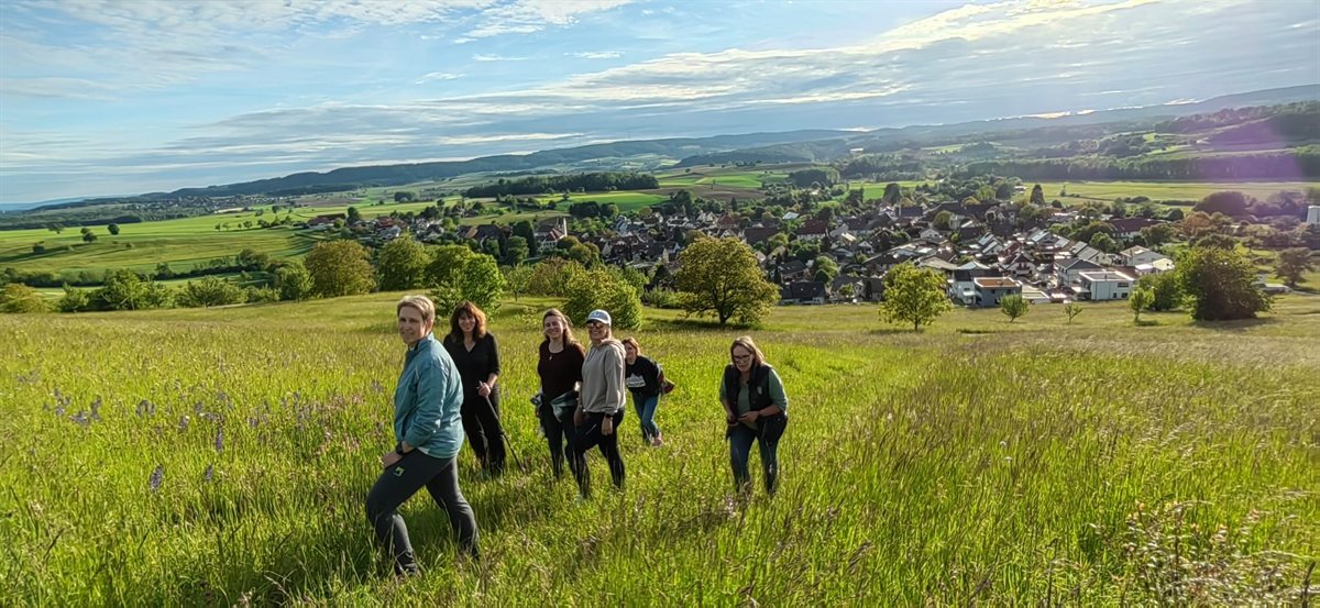 Ausflug der Fitnessdamen: Gruppe beim Wandern über eine Wiese mit Blick auf ein Dorf
