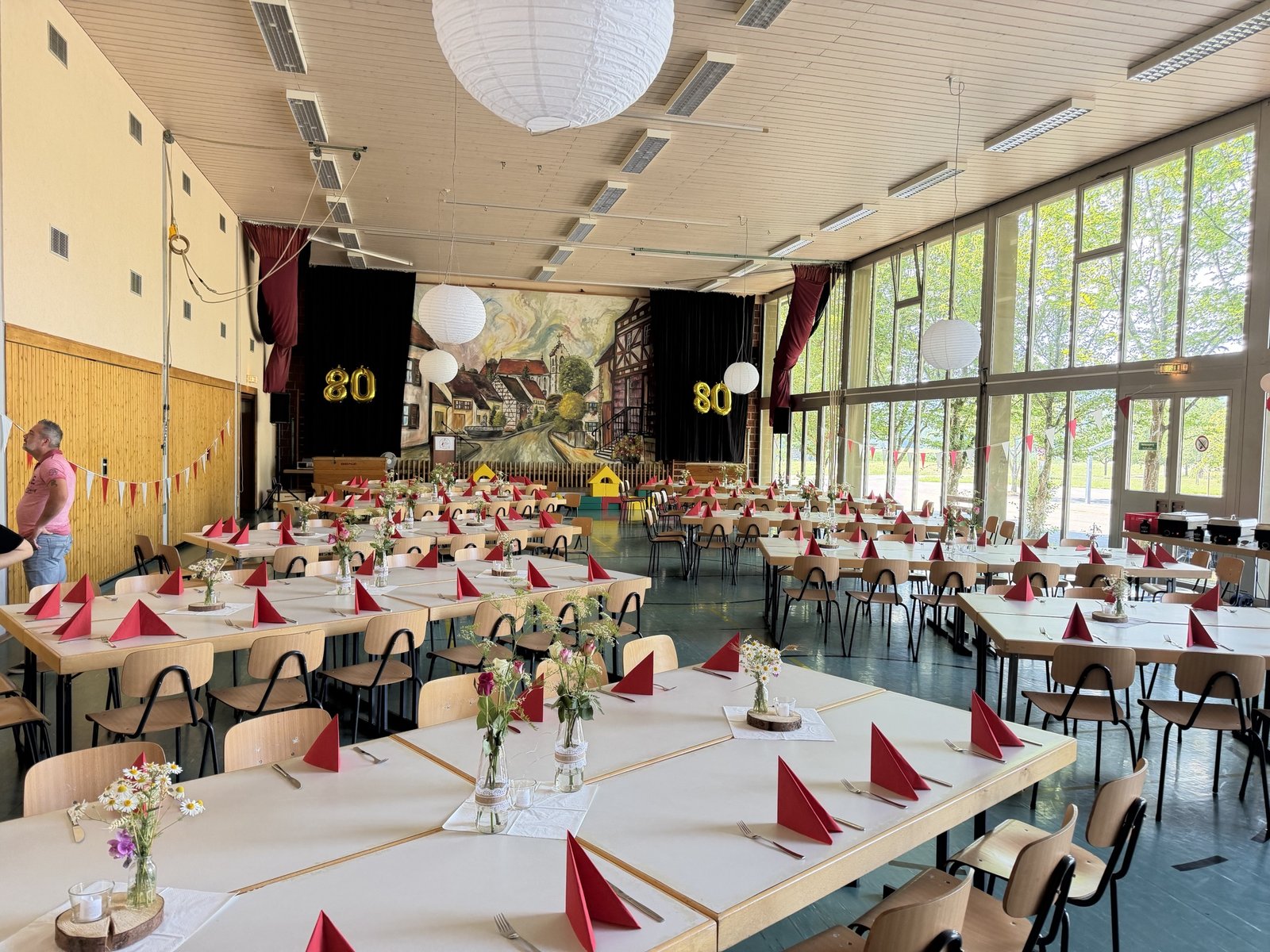 Festlich geschmückte Halle mit gedeckten Tischen, roten Servietten und goldenen Ballons „80“ vor der Bühne